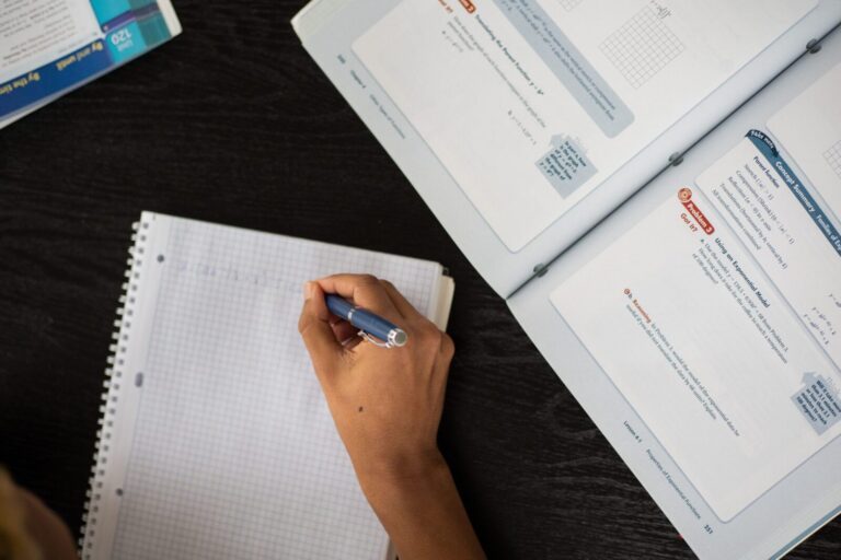 Top view of anonymous African American student taking notes in workbook at table with textbook while studying in classroom during lesson